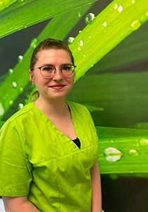 Eine freundlich lächelnde Frau mit Brille und hochgestecktem braunem Haar blickt in die Kamera. Sie trägt ein hellgrünes Praxisshirt. Im unscharfen Hintergrund ist ein grünes Naturmotiv aus Grashalmen mit Wassertropfen zu sehen.