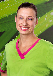 Ein lächelndes weibliches Teammitglied der Zahnarztpraxis Finger, gekleidet in grüne Arbeitskleidung mit rosa Akzenten, vor einem üppig grünen, blätterreichen Hintergrund. Ihr dunkles Haar ist zurückgebunden.