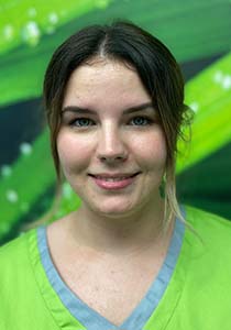 Ein Porträt einer lächelnden jungen Frau mit dunklem Haar und hellen Augen, die grüne medizinische Kleidung trägt. Sie steht vor einem verschwommenen grünen Hintergrund. Dies ist Sabrina, ein Mitglied des Teams der Zahnarztpraxis Finger.